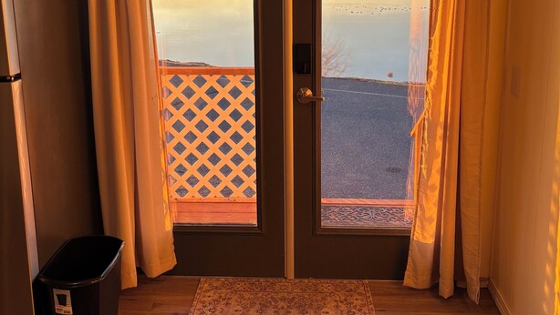 A room with a wooden floor, a black trash bin, a patterned rug, and a view of a calm body of water during sunset. - Sunset Paradise (Moses Lake)