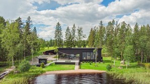Une maison moderne dotée d’une grande baie vitrée, entourée d’une forêt et bordée par un plan d’eau.
