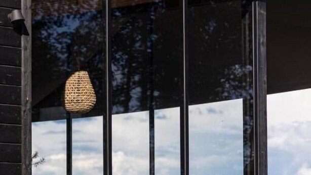 Une cabane moderne en bois, dotée d’un balcon, de grandes fenêtres et d’un luminaire tressé.