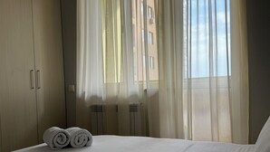 A hotel room with a bed, a window with sheer curtains, and a view of buildings outside.