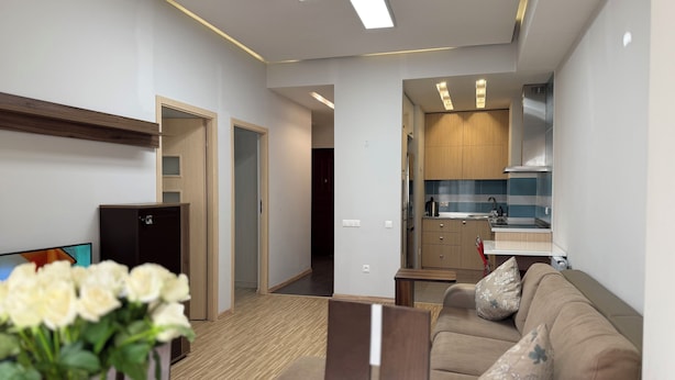 A modern living room with a sofa, a coffee table, and a TV. There is a kitchen area with cabinets and a stove.