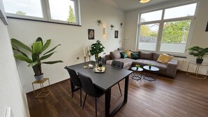 A modern living room with a dark wood floor, a grey sofa with cushions, a dining table with chairs, and large windows providing natural light. - Kings & Queens - 2 bedrooms - whirlpool - garden - barbecue - parking lot (Dudenhofen)