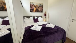 A neatly made bed with a purple blanket and two folded towels, a grey headboard, white pillows, and a painting of a Highland cow above the bed. - Kings & Queens - 2 bedrooms - whirlpool - garden - barbecue - parking lot (Dudenhofen)
