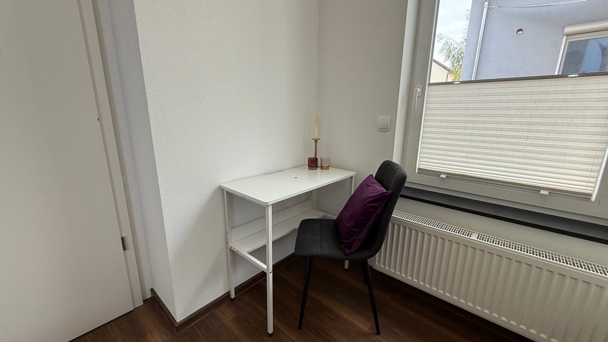 A small, white desk with a chair, a window with blinds, and a radiator.