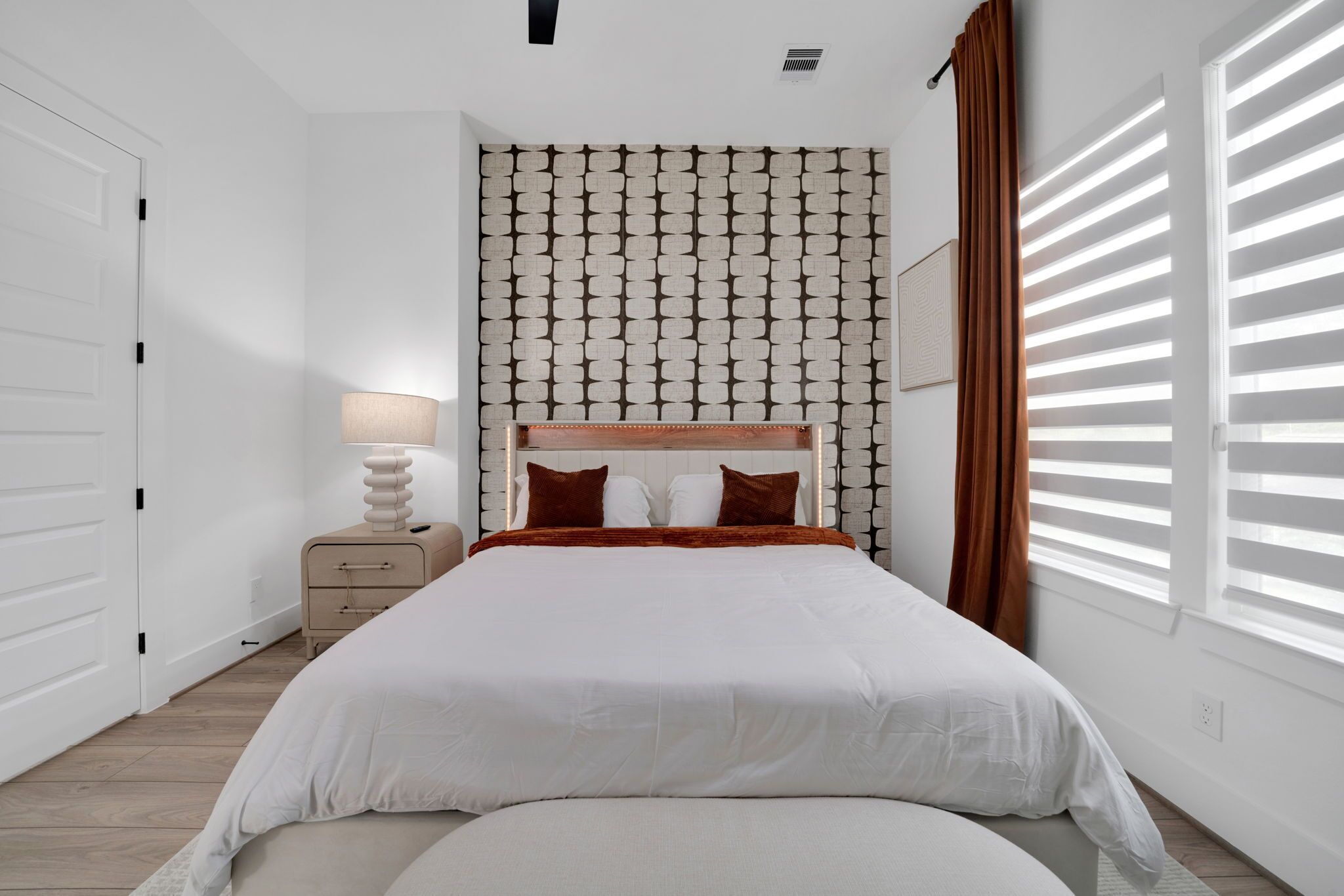 Une chambre à coucher avec un lit, une table de chevet, une lampe, un mur à motifs et une fenêtre avec des stores.