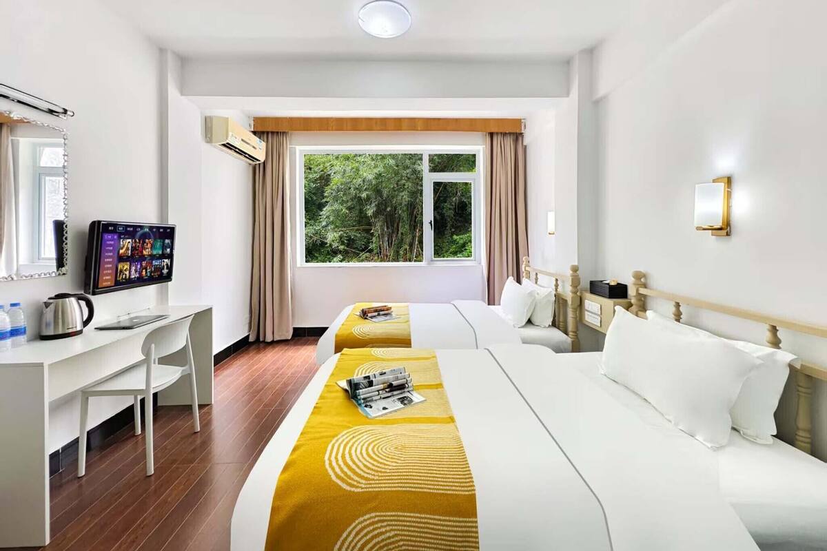 A hotel room with two beds, a desk, a television, and a window with a view of greenery.