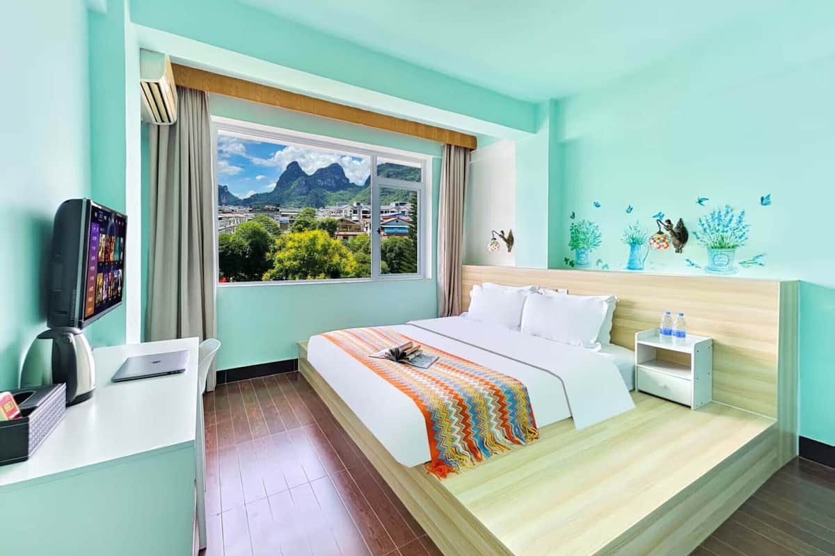A hotel room with a bed, a desk, a TV, and a window with a view of a mountain and town.