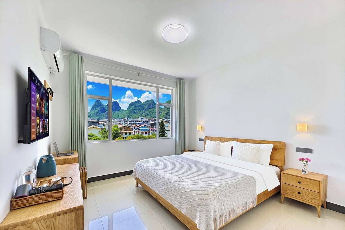 A hotel room with a large bed, a desk with a computer, a TV mounted on the wall, and a window with a city and mountains view.
