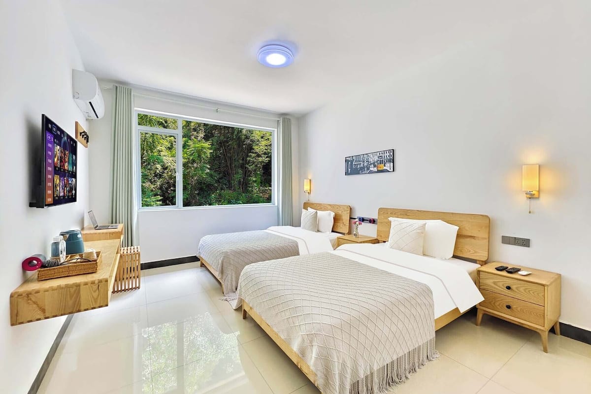 A modern hotel room with two beds, a large window, a flat-screen TV, and a small wooden side table.