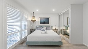 Ein modernes Schlafzimmer mit einem großen Bett, einer Pendelleuchte, einem gerahmten Bild an der Wand und einem Spiegel.