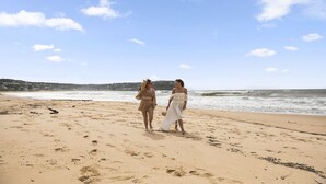 Zwei Personen laufen am sandigen Strand, im Hintergrund das Meer mit Wellen.