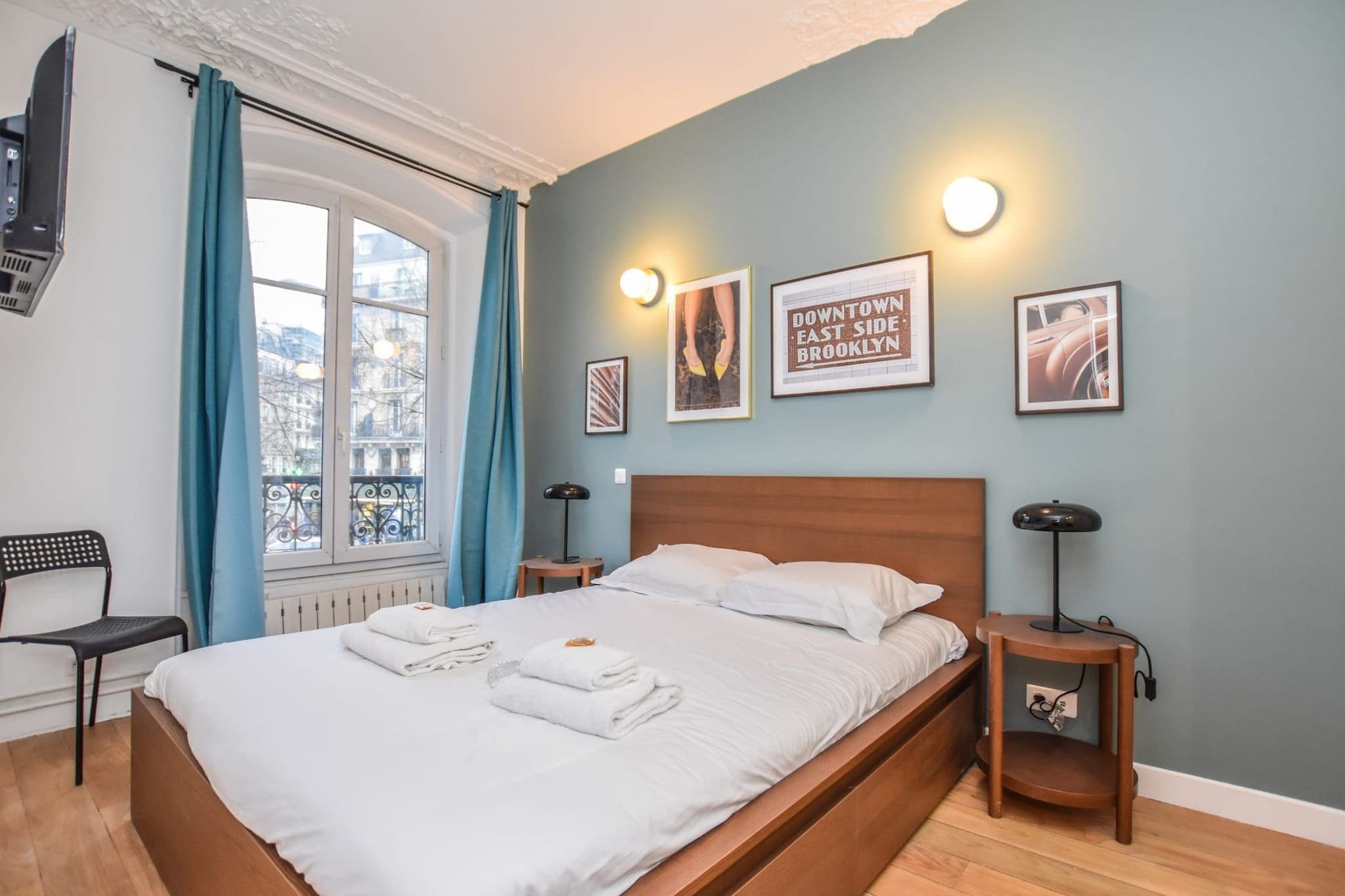 A neatly made bed with white linens and towels, a wooden headboard, a nightstand with a lamp, and a window with blue curtains.