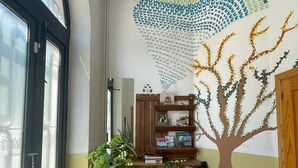 A room with a large window, a wooden desk with a potted plant, a bookshelf, and a wall mural of a tree. - El Vagón Bar-Albergue Béjar (Béjar)