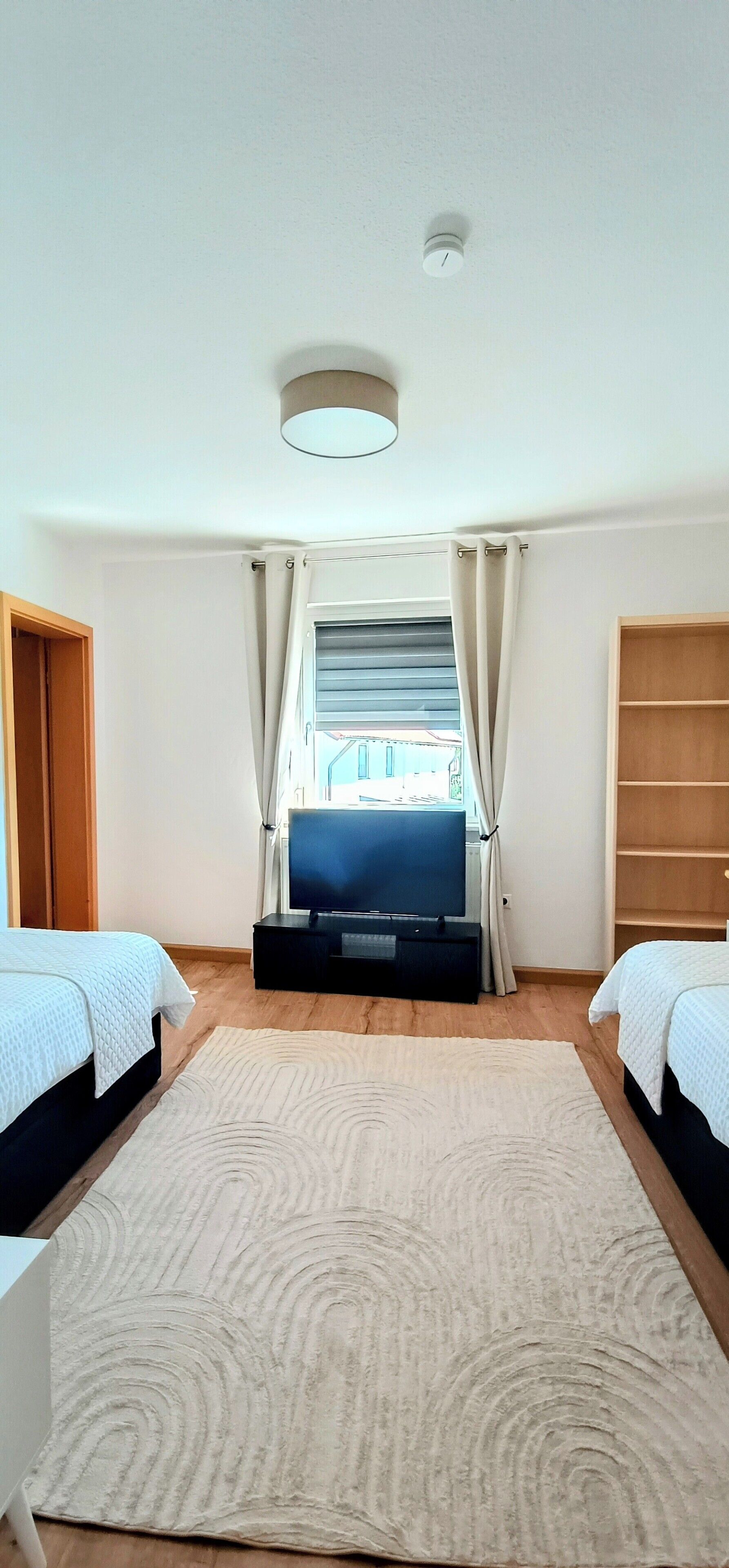Een slaapkamer met twee bedden, een televisie, een plafondlamp en een raam met gordijnen.