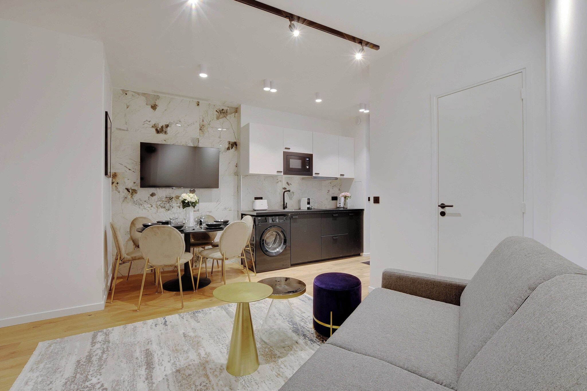 A modern living room with a grey sofa, a dining table with chairs, a TV mounted on the wall, and a kitchen area with a washing machine.