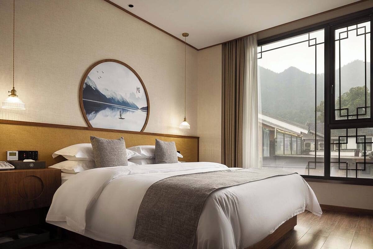 A hotel room with a large bed, a nightstand with a phone, a wall-mounted picture, and a view of a mountain and train tracks through a window.