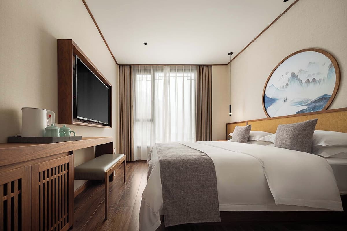 A hotel room with a bed, a desk with a television, a painting on the wall, and a window with curtains.