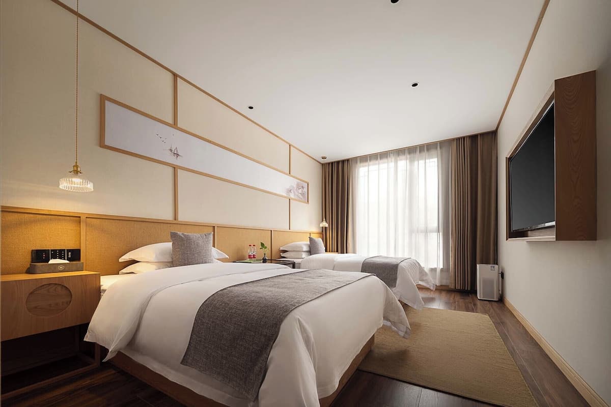 A hotel room with two beds, a flat-screen TV, a wooden headboard, a bedside table with a lamp, and a large window with curtains.