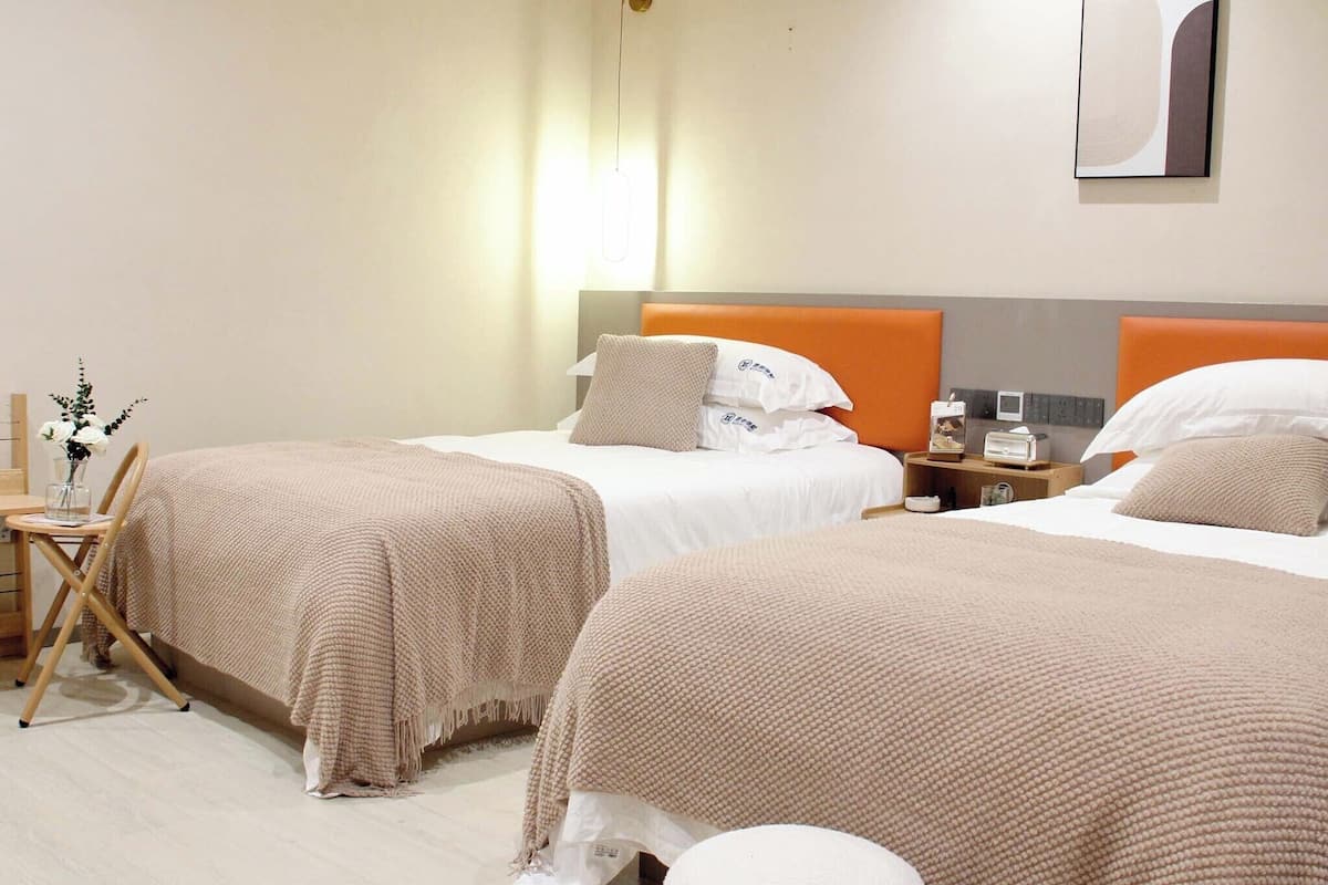 A hotel room with two beds, each with a beige blanket and pillows, a bedside table with a lamp, and a wall-mounted artwork.