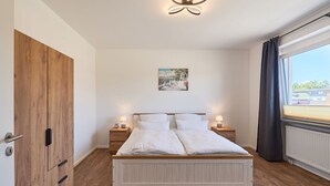 A bedroom with a wooden wardrobe, a bed with white bedding, two bedside tables with lamps, a wall-mounted picture, and a window with curtains.