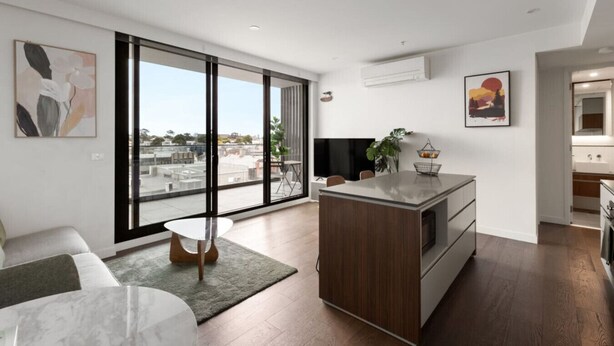 A modern apartment with a large sliding glass door, a kitchen with a central island, a dining area, and a living room with a large window offering a city view. - Stylish apartment near South Melbourne Market (South Melbourne)