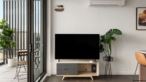 A modern living room with a large flat-screen TV on a wooden TV stand, a dining area with a table and chairs, and a balcony with potted plants. - Stylish apartment near South Melbourne Market (South Melbourne)