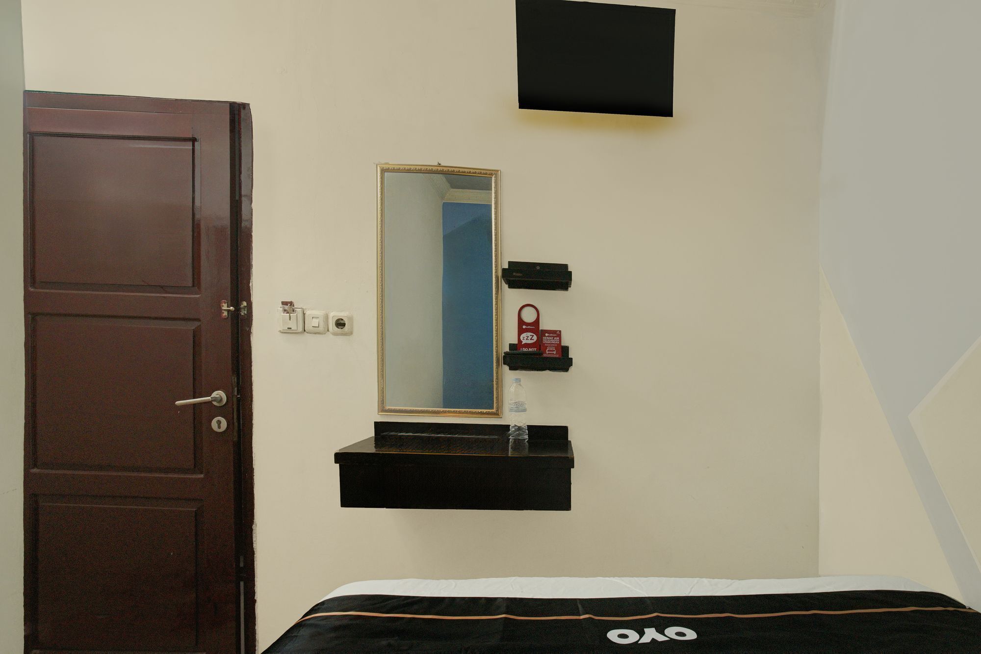 Una habitación con cama, puerta, espejo y un pequeño estante con objetos.