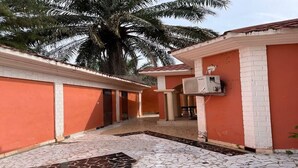 A covered outdoor area with tiled flooring, a palm tree, and a building with a red and white facade. - Serene Room and Living Room (Serrekunda)