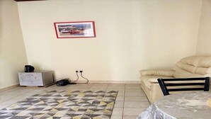 A room with a beige sofa, a small table with a plastic cover, a patterned rug, a framed picture on the wall, and a cabinet with a coffee maker. - Serene Room and Living Room (Serrekunda)
