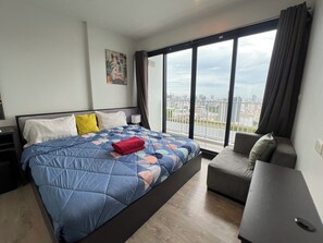A modern bedroom with a large bed, a grey sofa, and a city view through floor-to-ceiling windows. - Exclusive Sea View 2 Bed Room High Floor 2 Balcony (Muang Pattaya)