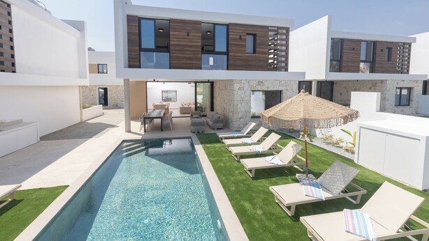 Una casa moderna de dos pisos con piscina, mobiliario exterior y un muro de piedra.