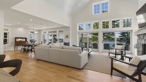 A spacious living area with a large sofa, dining table, and chairs, a kitchen area, and a view of the outdoors through large windows. - Ocean Point Boothbay Luxury Home|5BR |4.5BA|Sleeps 12 (Boothbay)