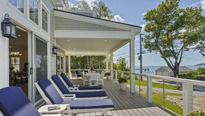 A covered outdoor deck with lounge chairs and a dining area, overlooking a body of water. - Ocean Point Boothbay Luxury Home|5BR |4.5BA|Sleeps 12 (Boothbay)