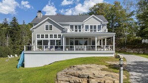 A two-story house with a covered porch, a stone retaining wall, and a grassy yard. - Ocean Point Boothbay Luxury Home|5BR |4.5BA|Sleeps 12 (Boothbay)