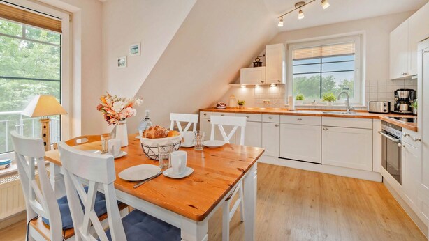 A modern kitchen with white cabinets, a wooden dining table, and a view of the outdoors through large windows.