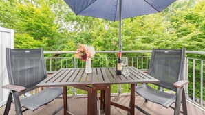 A wooden table and two chairs with a mesh backrest, a large umbrella, a vase with flowers, and a bottle of wine on the balcony.