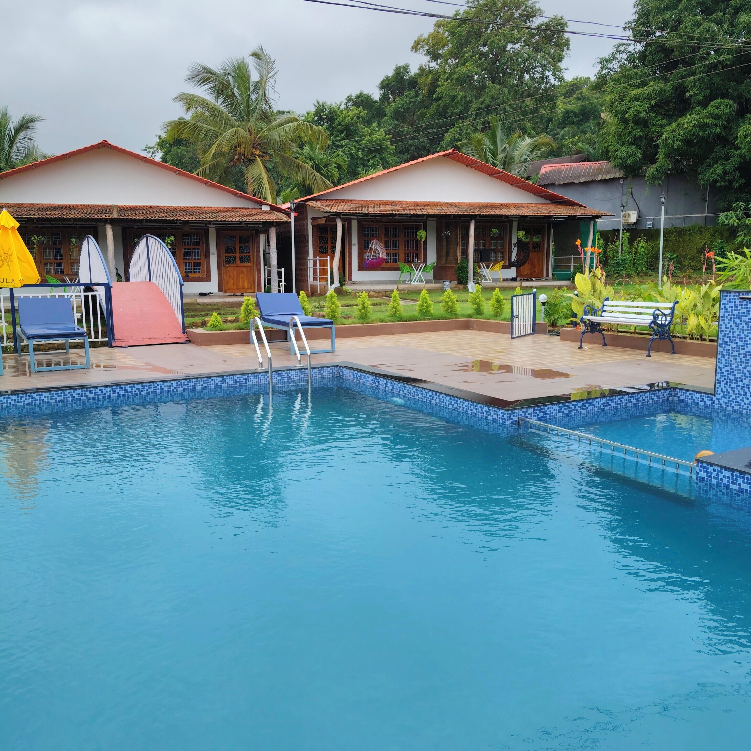 A swimming pool with lounge chairs and umbrellas, surrounded by a garden and a building with a tiled roof.
