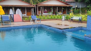 A swimming pool with lounge chairs and umbrellas, surrounded by a garden and a building with a tiled roof.