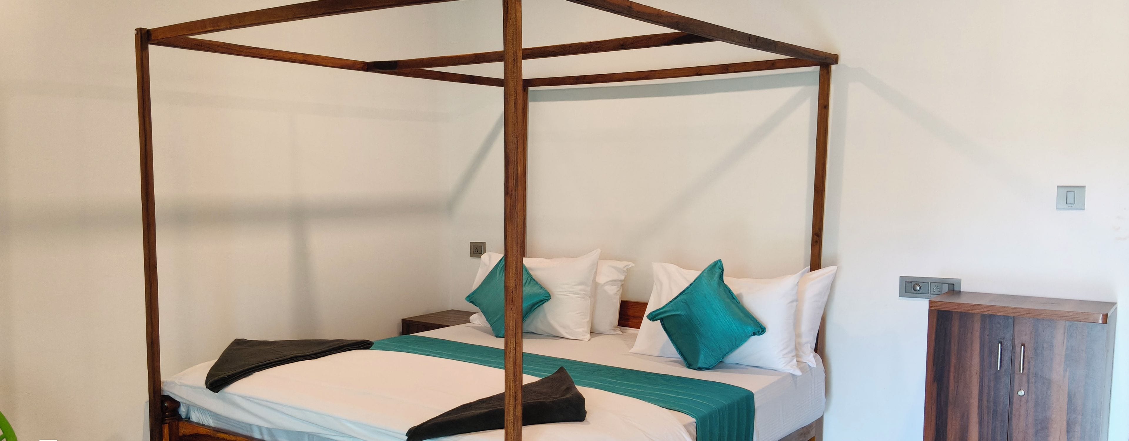 A four-poster bed with white linens and teal pillows, a wooden nightstand, and a wardrobe with a dark finish.