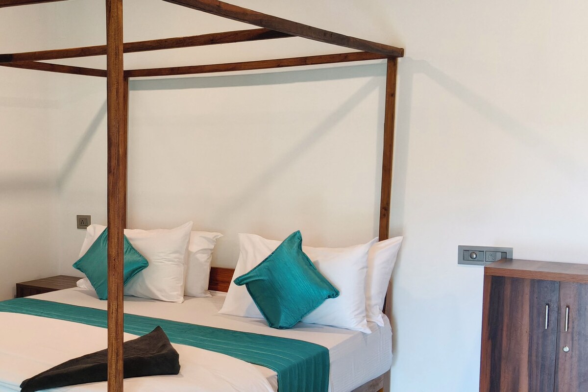 A four-poster bed with white linens and teal pillows, a wooden nightstand, and a wardrobe with a dark finish.
