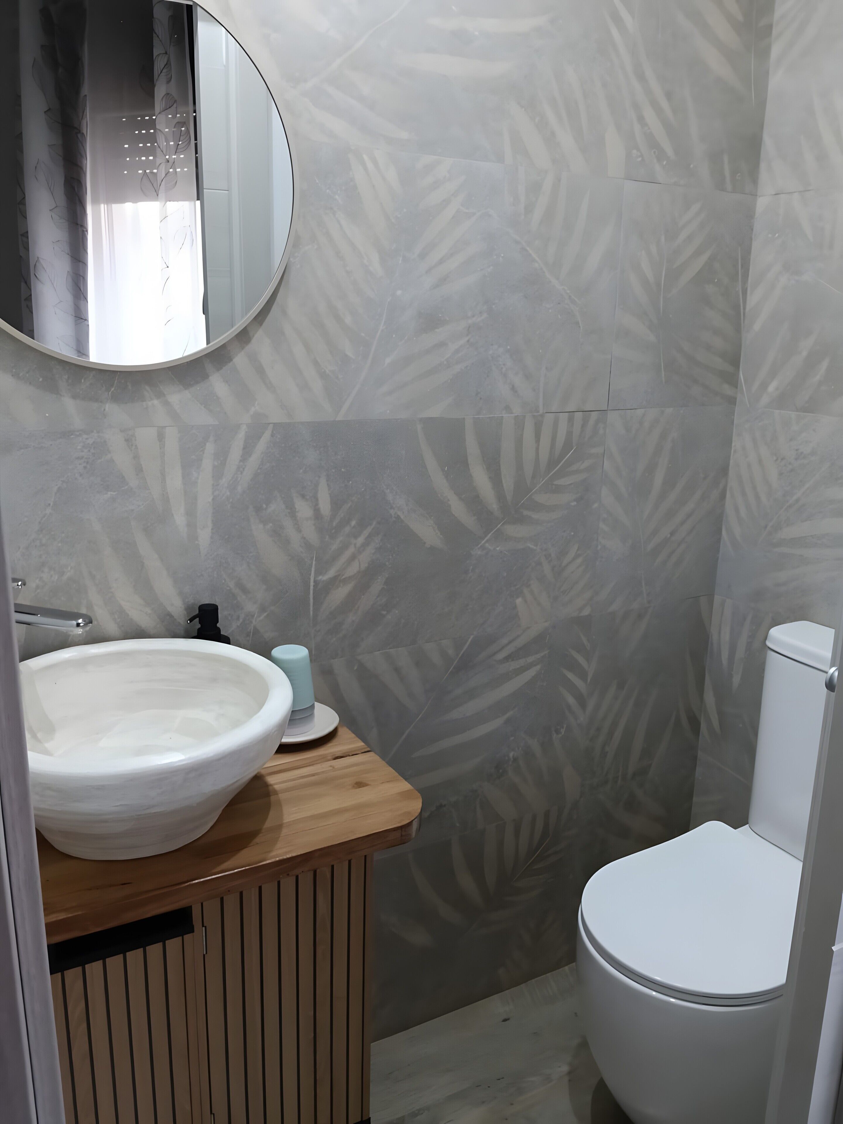 Un baño con un espejo redondo, un lavamanos blanco, un inodoro y baldosas estampadas.