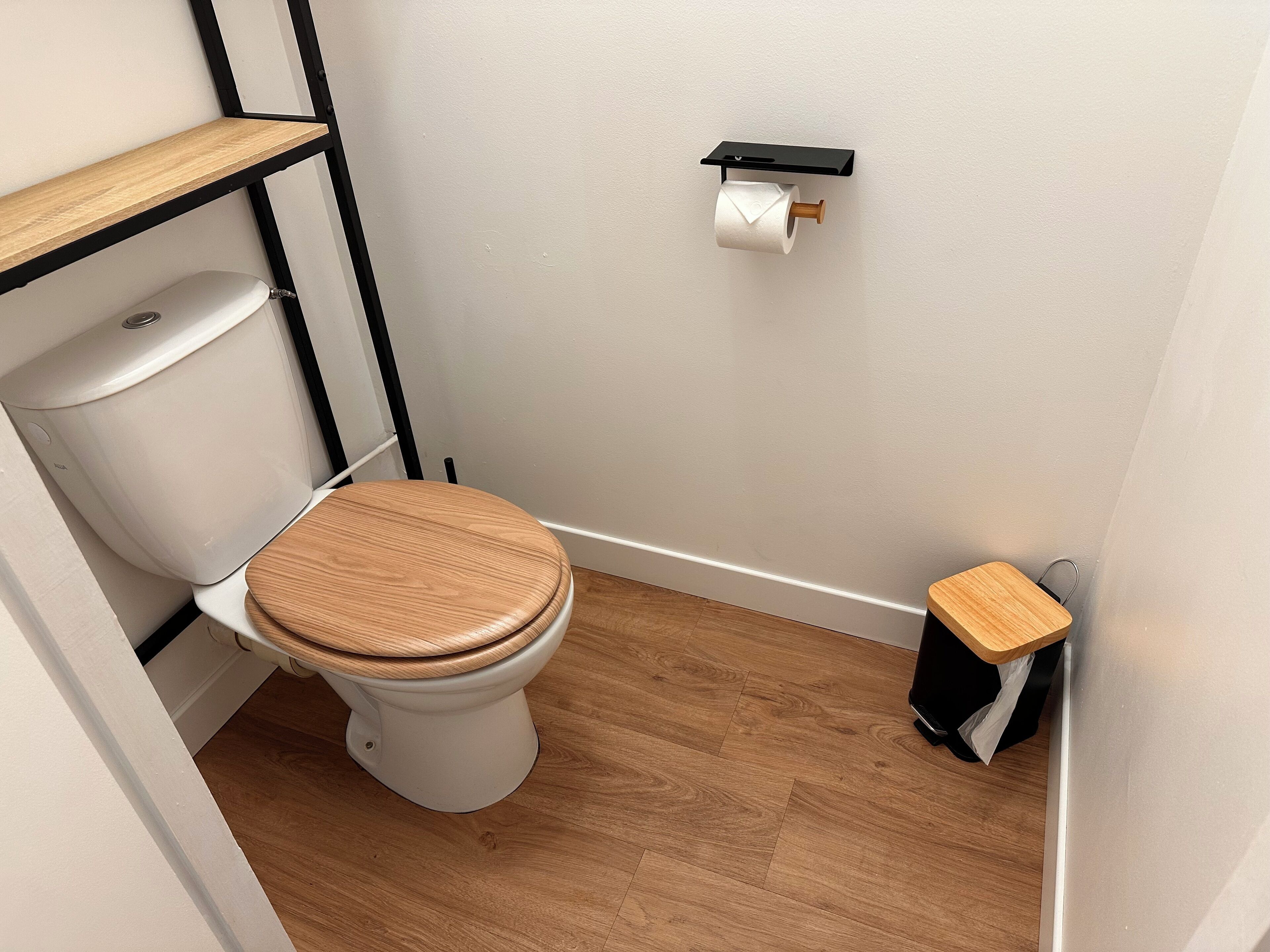 Een kleine, schone badkamer met toilet, houten vloer en een plank boven het toilet.