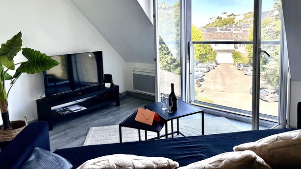 A modern living room with a large TV, a dining table with a bottle and glasses, and a view of a parking lot through large windows. - Le Ronsard, Chinon View of the fortress & Collegiate church of Saint Mexme - Historic center 5mn (Chinon)