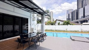 Un patio moderno al aire libre con una mesa y sillas de vidrio, una piscina y edificios altos al fondo.