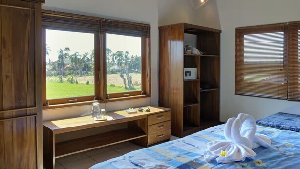 A bedroom with a wooden desk, a bed with a blue bedspread, and a view of palm trees through a window. - The Studio at Warung Bambu Pemaron (Buleleng)