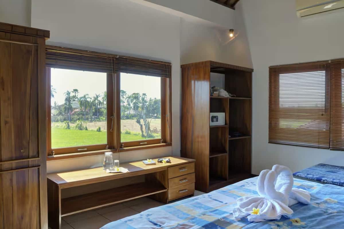 A bedroom with a wooden desk, a bed with a blue bedspread, and a view of palm trees through a window.