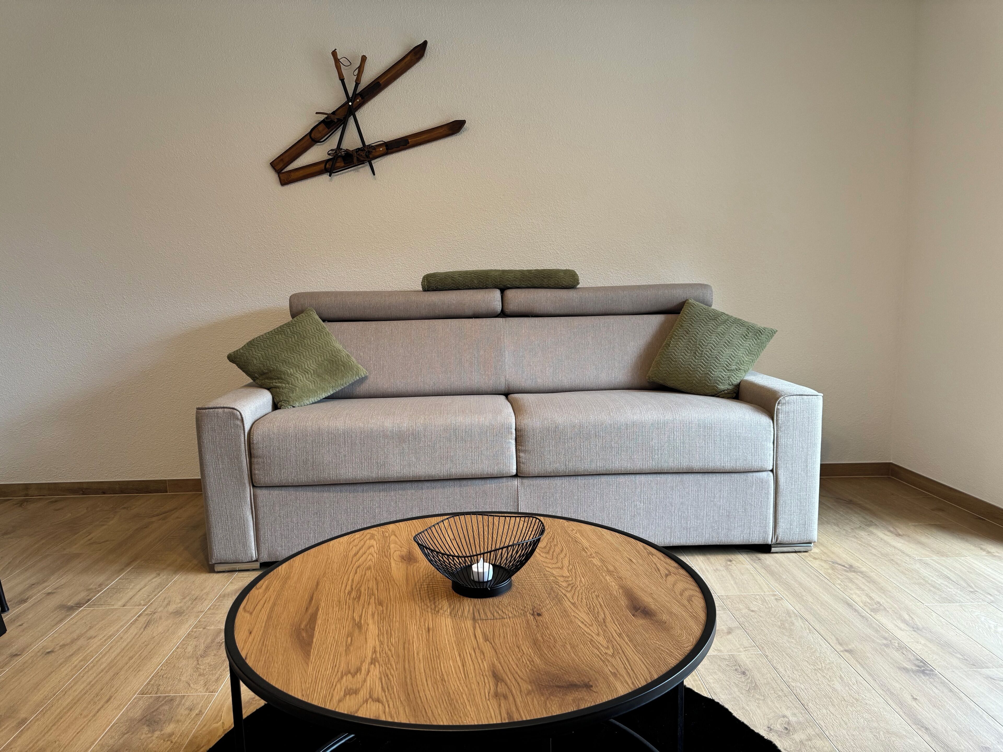 Un salon moderne avec un canapé gris, deux coussins verts et une table basse ronde en bois, dont le pied est en métal noir.