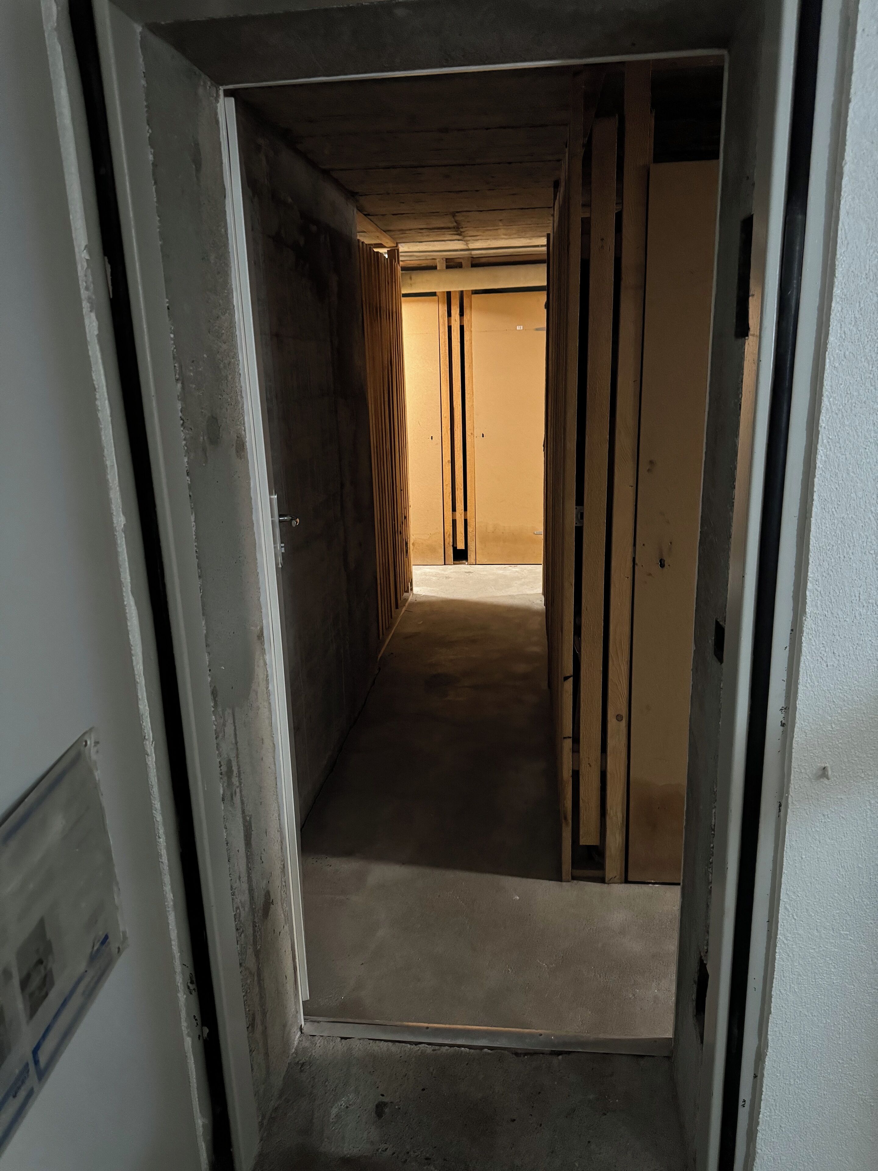 Um corredor estreito com vigas de madeira aparentes e chão de concreto, que leva a uma porta fechada no final.