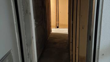 Um corredor estreito com vigas de madeira aparentes e chão de concreto, que leva a uma porta fechada no final.
