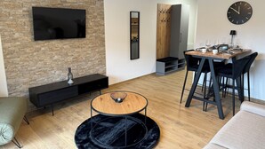 Una sala de estar moderna con una pared de piedra, un televisor de pantalla plana, una mesa de centro redonda, una zona de comedor con una mesa de madera y sillas negras, y un reloj negro en la pared.